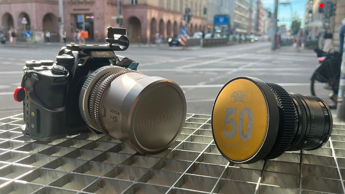 Two camera lenses resting on a metal grate with a blurred city intersection behind them; the left is a silver DZOFILM lens attached to a black camera cage, and the right is a black lens with a yellow Contax Zeiss 50 cap