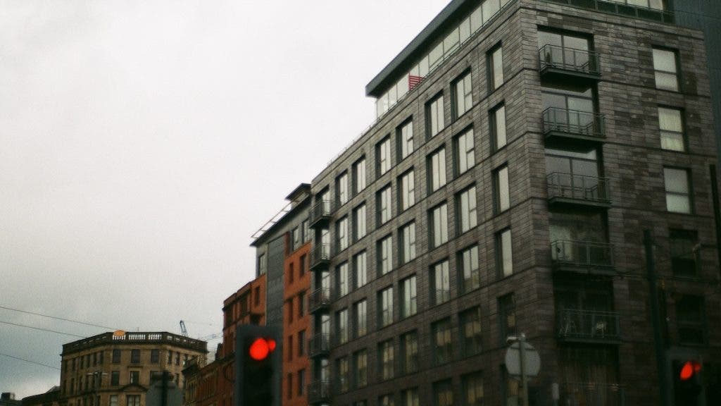 A street-level perspective of a modern, multi-story dark stone apartment building against an overcast sky, with red traffic lights glowing in the foreground