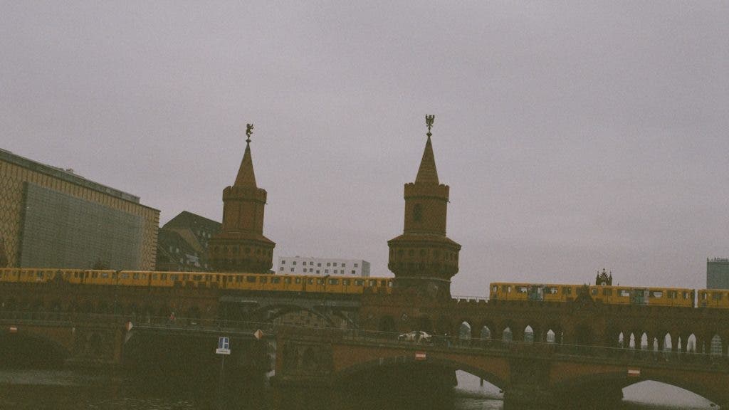 A wide, hazy view of the historic Oberbaum Bridge in Berlin with its twin spired towers, featuring a yellow U-Bahn train crossing the upper deck over the river