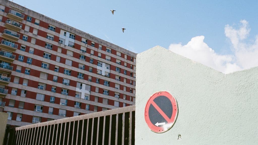 A low-angle shot of a pale green concrete wall featuring a circular red and blue "No Entry / No Parking" sign with a white arrow. In the background, a large multi-story residential building with a grid of windows sits under a bright blue sky with two birds flying above