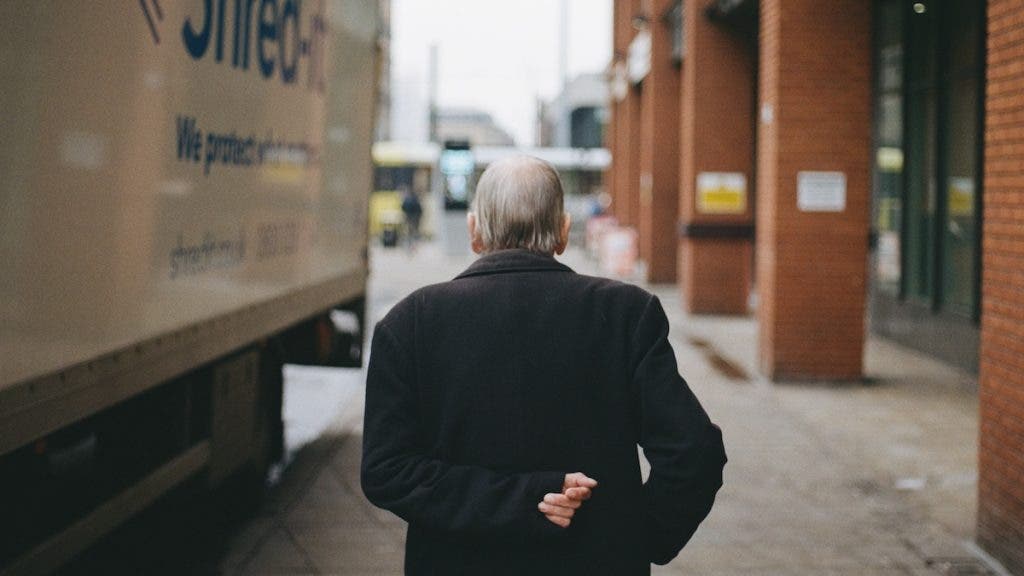 Back view of an elderly man in a dark coat walking down a city sidewalk past a parked Shred-it truck and brick building pillars