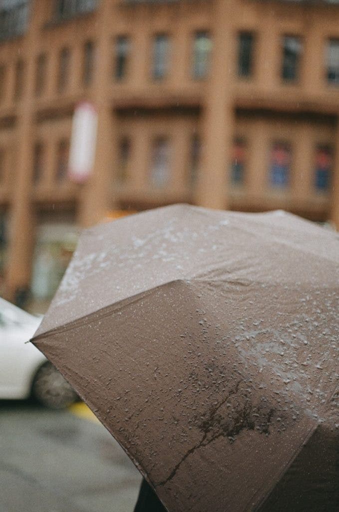 A close-up of a wet, brown umbrella covered in raindrops, with a softly blurred background showing a brick building and the edge of a white car