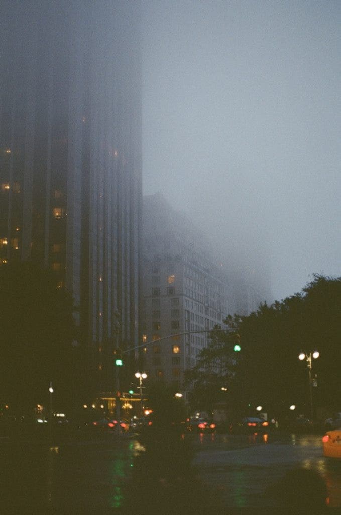 A foggy evening cityscape showing tall buildings partially obscured by heavy mist, with illuminated windows and glowing streetlights reflecting on a wet road