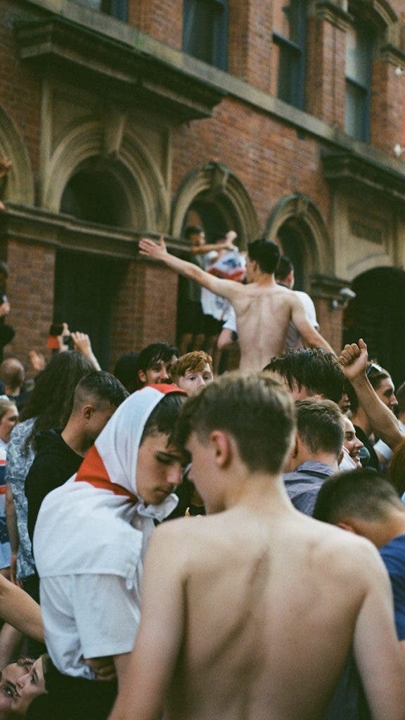A crowd of young supporters, some wearing St George's Cross flags and one shirtless with his arm raised, gathering outside a brick building during a celebratory outdoor event