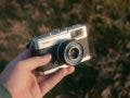A hand holding a vintage silver and black Olympus Trip 35 compact film camera against a softly blurred, warm outdoors background