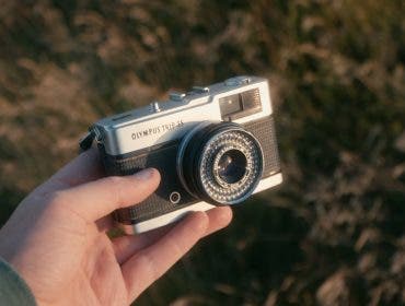A hand holding a vintage silver and black Olympus Trip 35 compact film camera against a softly blurred, warm outdoors background