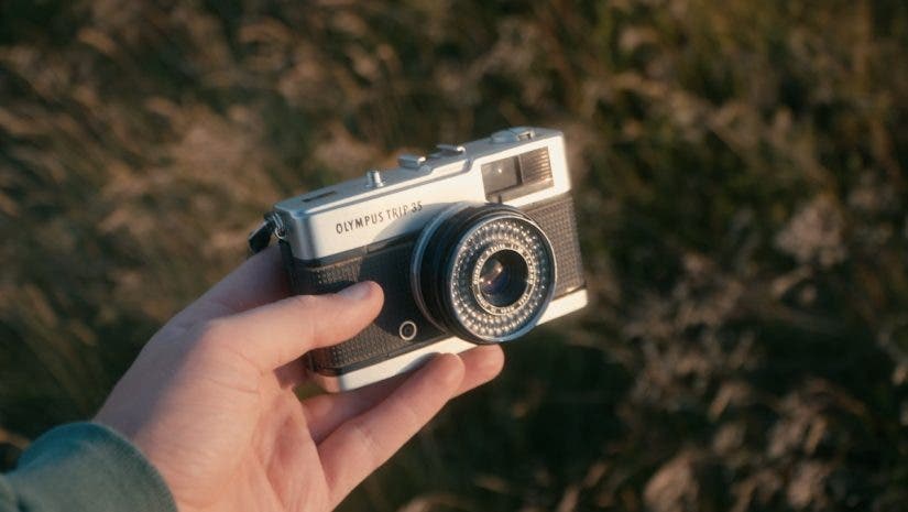 A hand holding a vintage silver and black Olympus Trip 35 compact film camera against a softly blurred, warm outdoors background