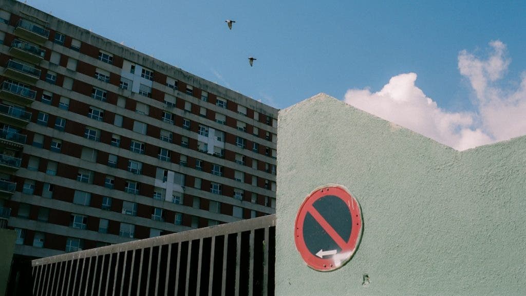 A low-angle shot of a pale green concrete wall featuring a circular red and blue "No Entry / No Parking" sign with a white arrow. In the background, a large multi-story residential building with a grid of windows sits under a bright blue sky with two birds flying above