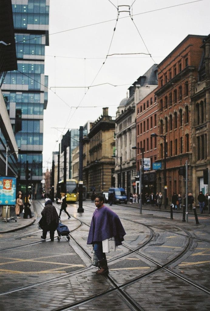 Pedestrians cross a cobblestone city street with curving tram tracks, featuring a woman in a purple poncho in the center foreground and a mix of modern and historical buildings lining the street under an overcast sky