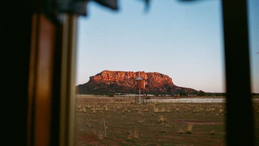 A framed view of a wide desert landscape featuring a small water tower and a red rock mesa mountain under a clear sky