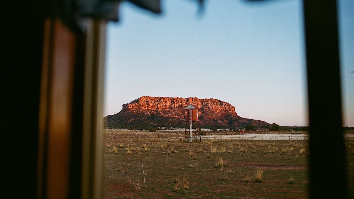 A framed view of a wide desert landscape featuring a small water tower and a red rock mesa mountain under a clear sky