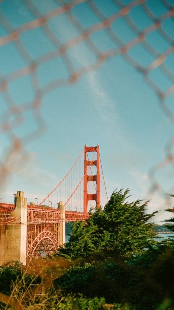 The Golden Gate Bridge in San Francisco framed through a blurred chain-link fence, surrounded by green trees under a clear blue sky