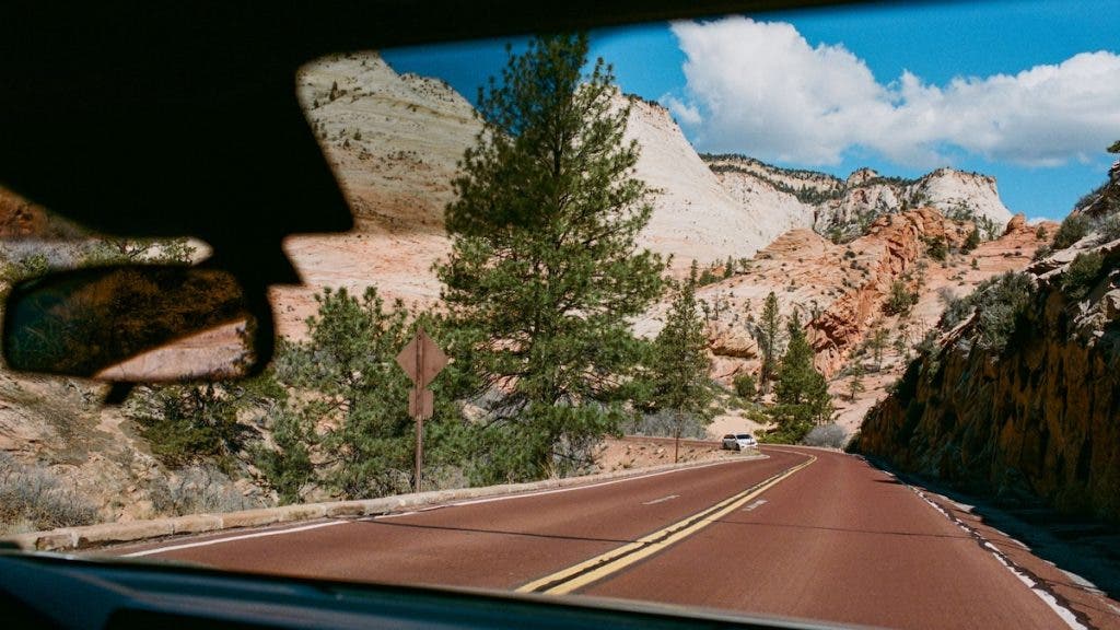 View from inside a moving vehicle framed by the windshield and rearview mirror, looking out onto a red paved winding road surrounded by pine trees and towering sandstone rock formations under a bright blue sky with light clouds