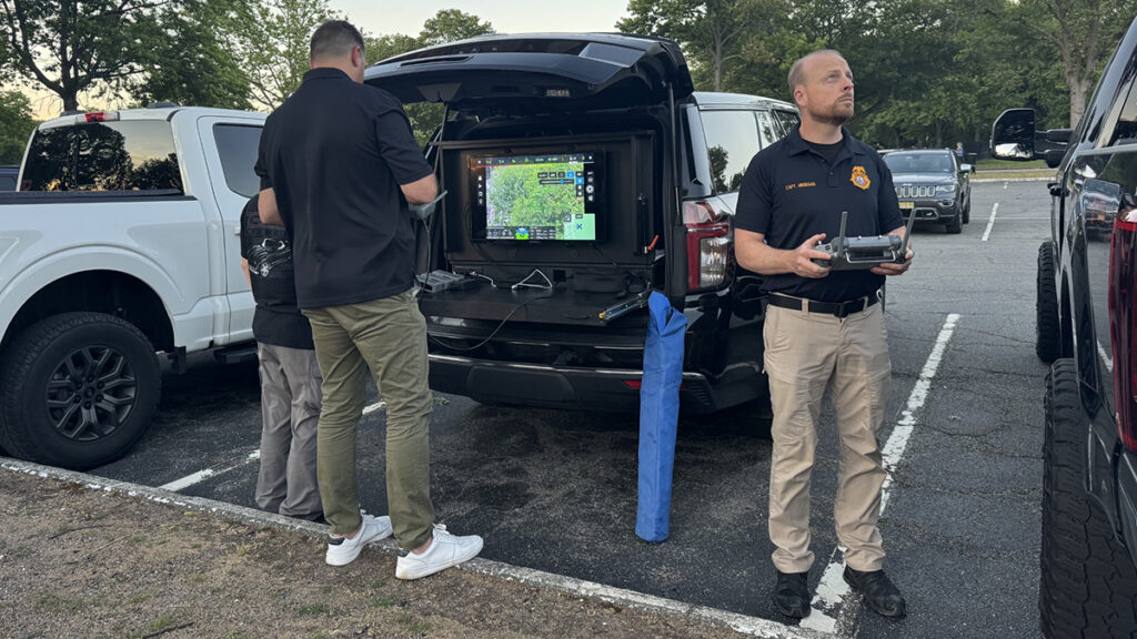 A mobile drone command station set up in a parking lot, featuring a large monitor mounted inside the open trunk of an SUV displaying flight telemetry, while a pilot looks up at the sky holding a remote control.